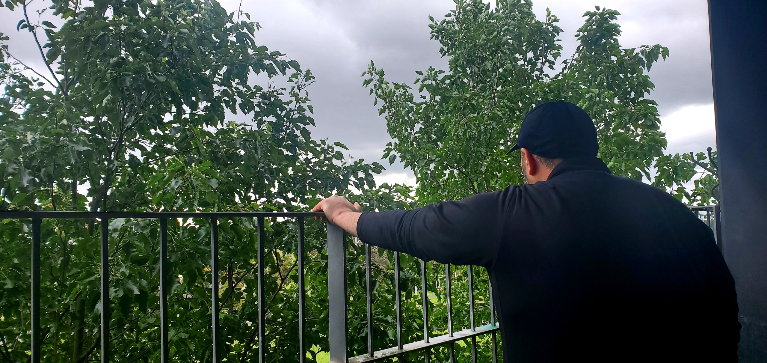 A person in a black jacket and cap stands on a balcony, facing away and looking at green trees under a cloudy, overcast sky.