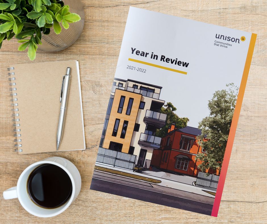 A Year in Review 2021-2022 booklet with an illustrated building on the cover lies on a wooden table next to a spiral notebook with a pen, a cup of black coffee, and a leafy plant.