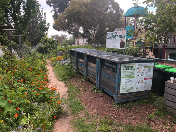 A community compost hub with large wooden compost bins beside a garden path, surrounded by lush greenery and flowers. Signs display compost hub information and rules.