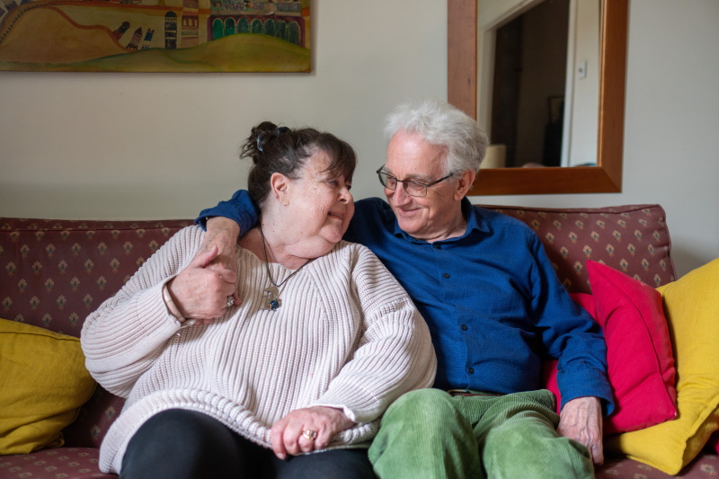 An older couple sits close together on a patterned couch, smiling warmly at each other. The man has his arm around the woman. Colorful cushions and a painting decorate the cozy living room.