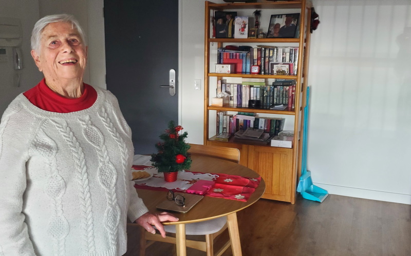 An elderly woman in a white sweater smiles while standing next to a small table decorated with a mini Christmas tree and placemats. A bookshelf and cleaning tools are visible in the background.