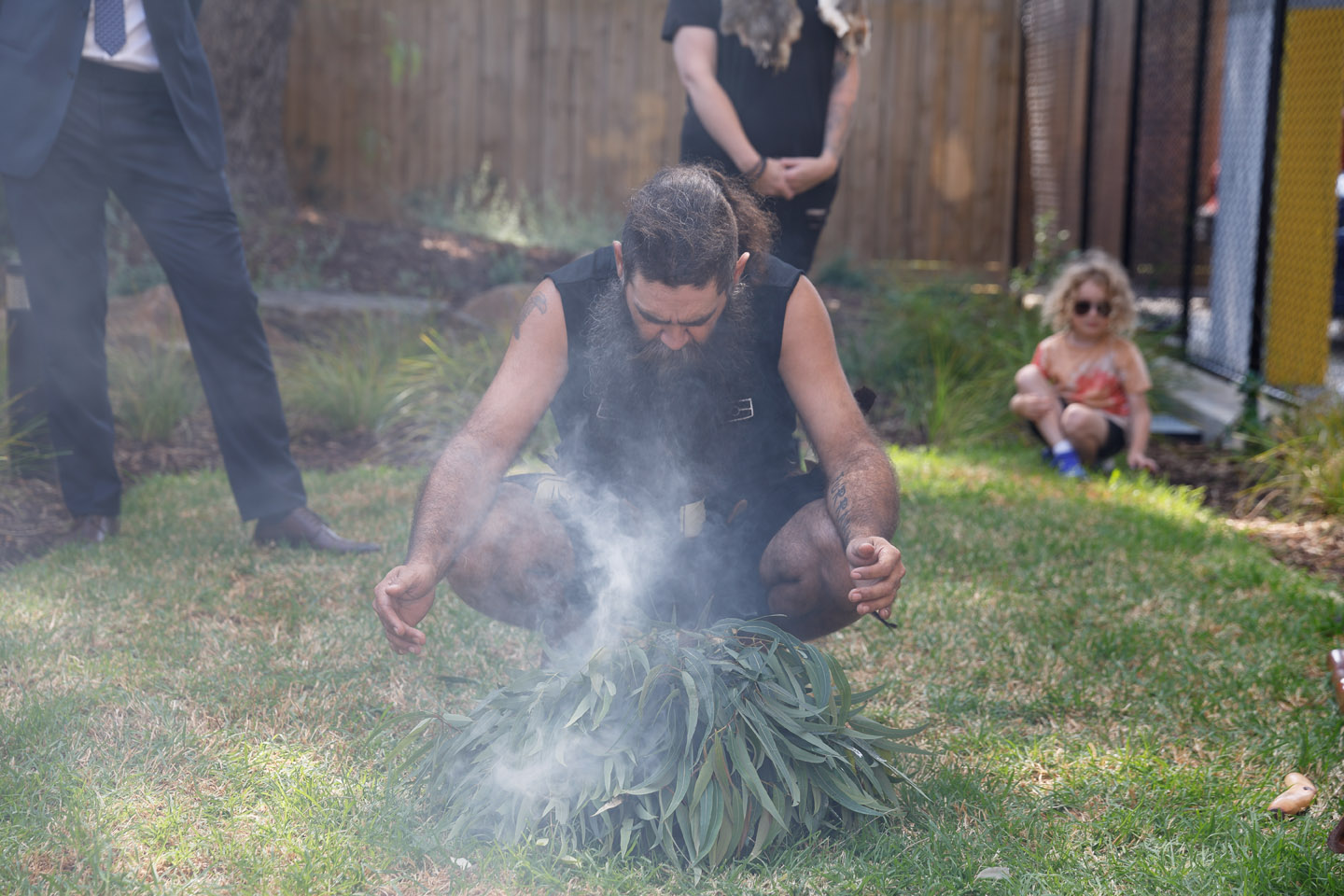 A man crouches over a smoking pile of eucalyptus leaves during a traditional ceremony outdoors, while people stand and sit nearby, including a child in the background.