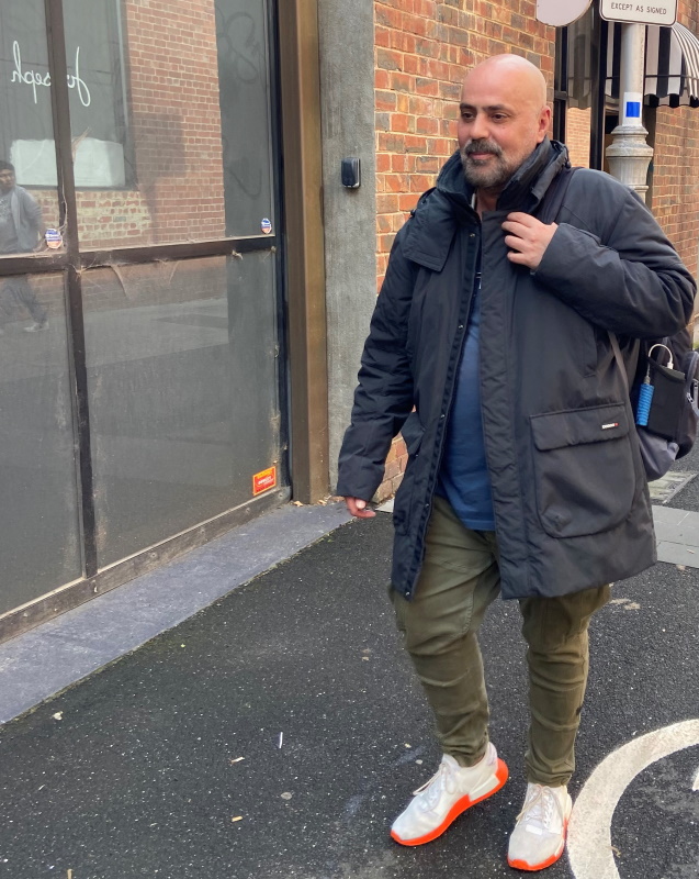 A man with a beard, wearing a dark coat, olive pants, and white sneakers with orange soles, walks on a wet city sidewalk near a brick building. He has a backpack over one shoulder.