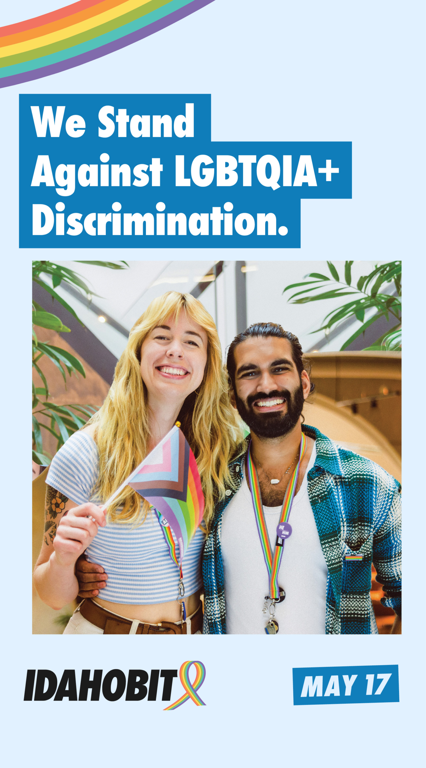 Two smiling people stand together, one holding a rainbow flag and both wearing rainbow lanyards. Text reads: We Stand Against LGBTQIA+ Discrimination. Below, IDAHOBIT and May 17 with a rainbow ribbon are shown.