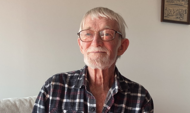 An elderly man with white hair, a beard, and glasses sits on a light-colored couch, wearing a black and white plaid shirt, smiling softly. A framed picture hangs on the wall behind him.