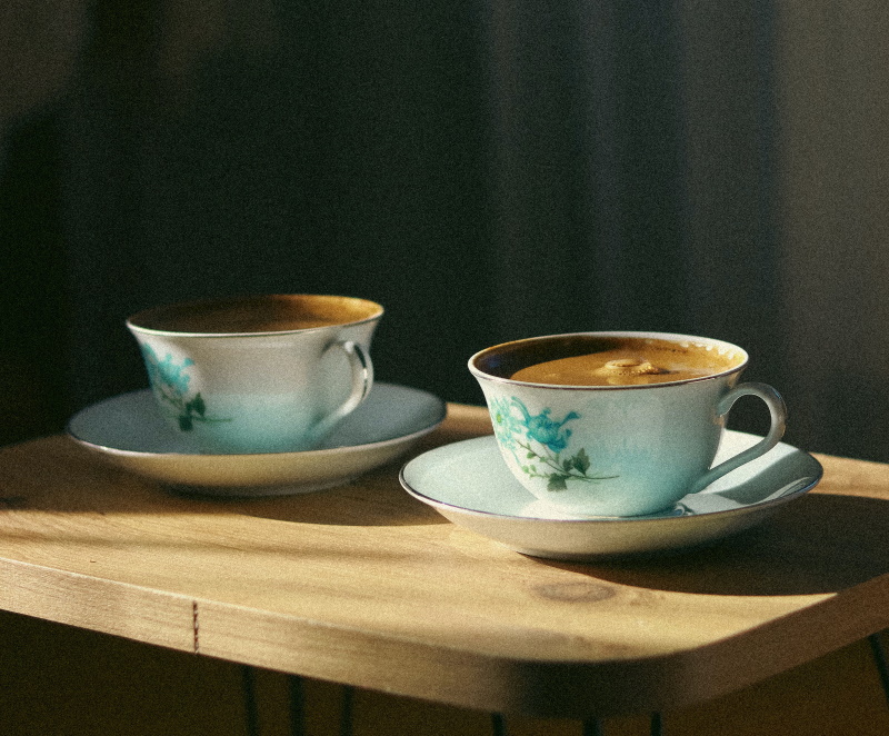 Two blue floral teacups filled with coffee sit on matching saucers atop a wooden table, with warm sunlight casting soft shadows across the scene.