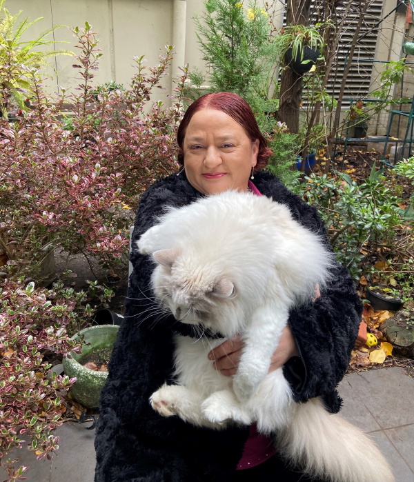 A woman with reddish-brown hair wearing a black coat stands outdoors, smiling and holding a large, fluffy white cat in her arms. Lush plants and greenery surround them in the background.