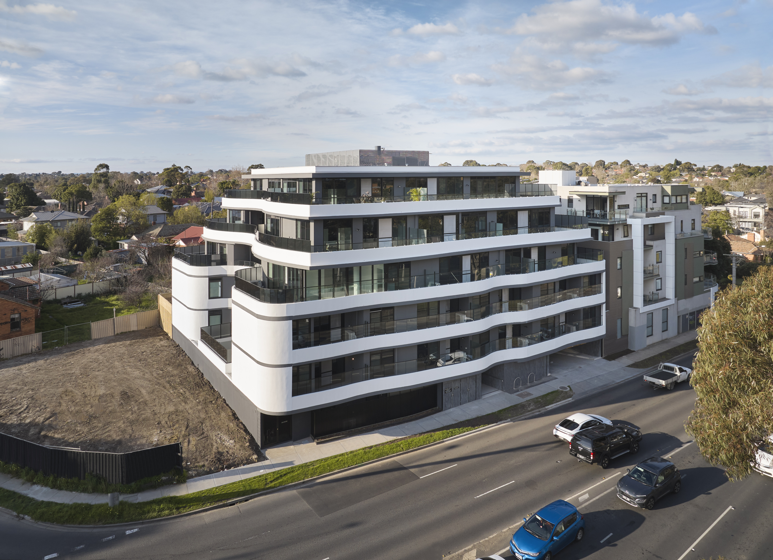 A modern, multi-story white apartment building with curved balconies stands beside a busy road with several cars. The building is surrounded by residential houses and an empty lot is visible on one side.