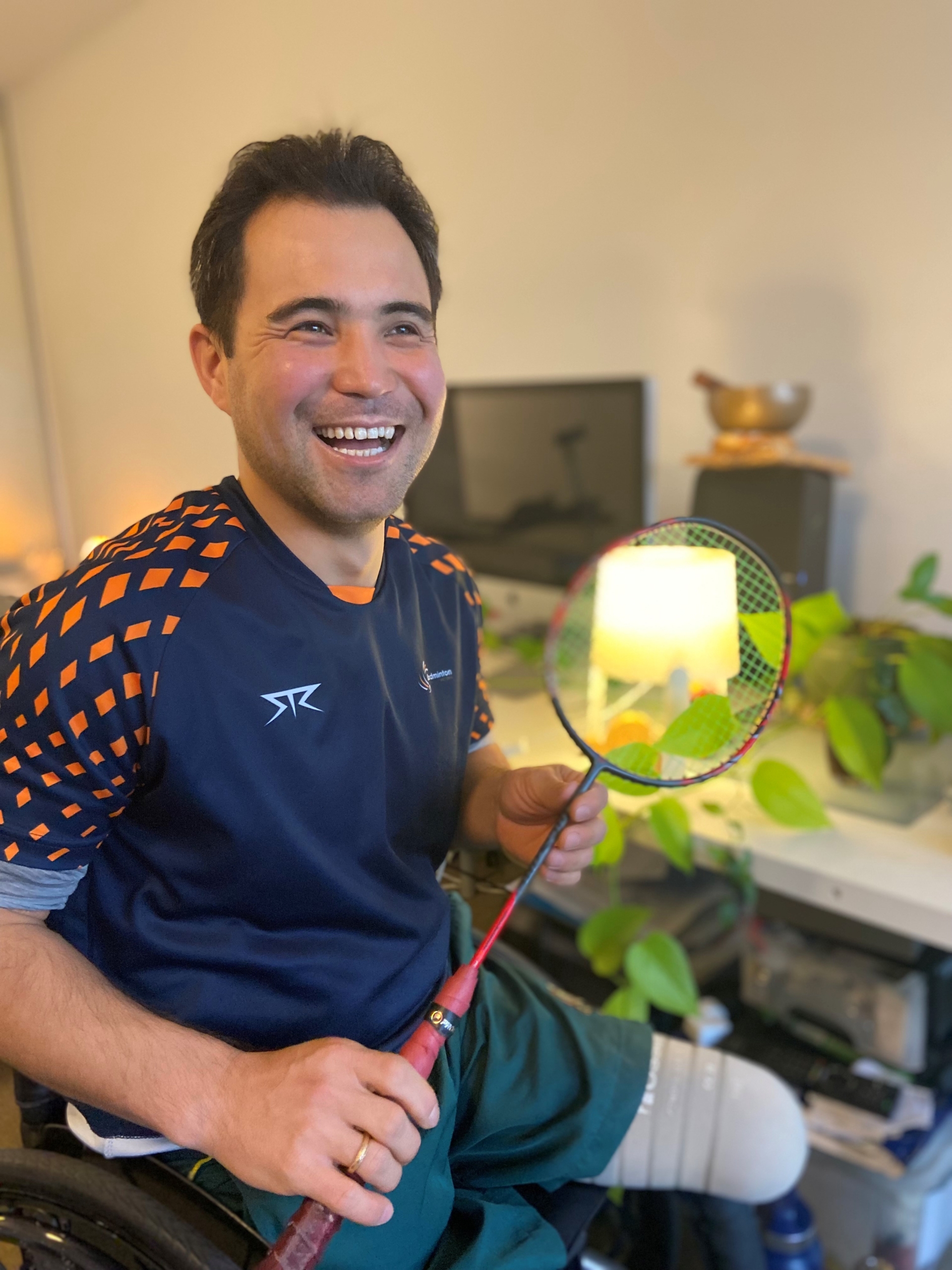 A smiling man in a sports wheelchair holds a badminton racket. He wears a navy sports shirt with orange accents and green shorts. A prosthetic leg is visible, and there are plants and a computer in the background.