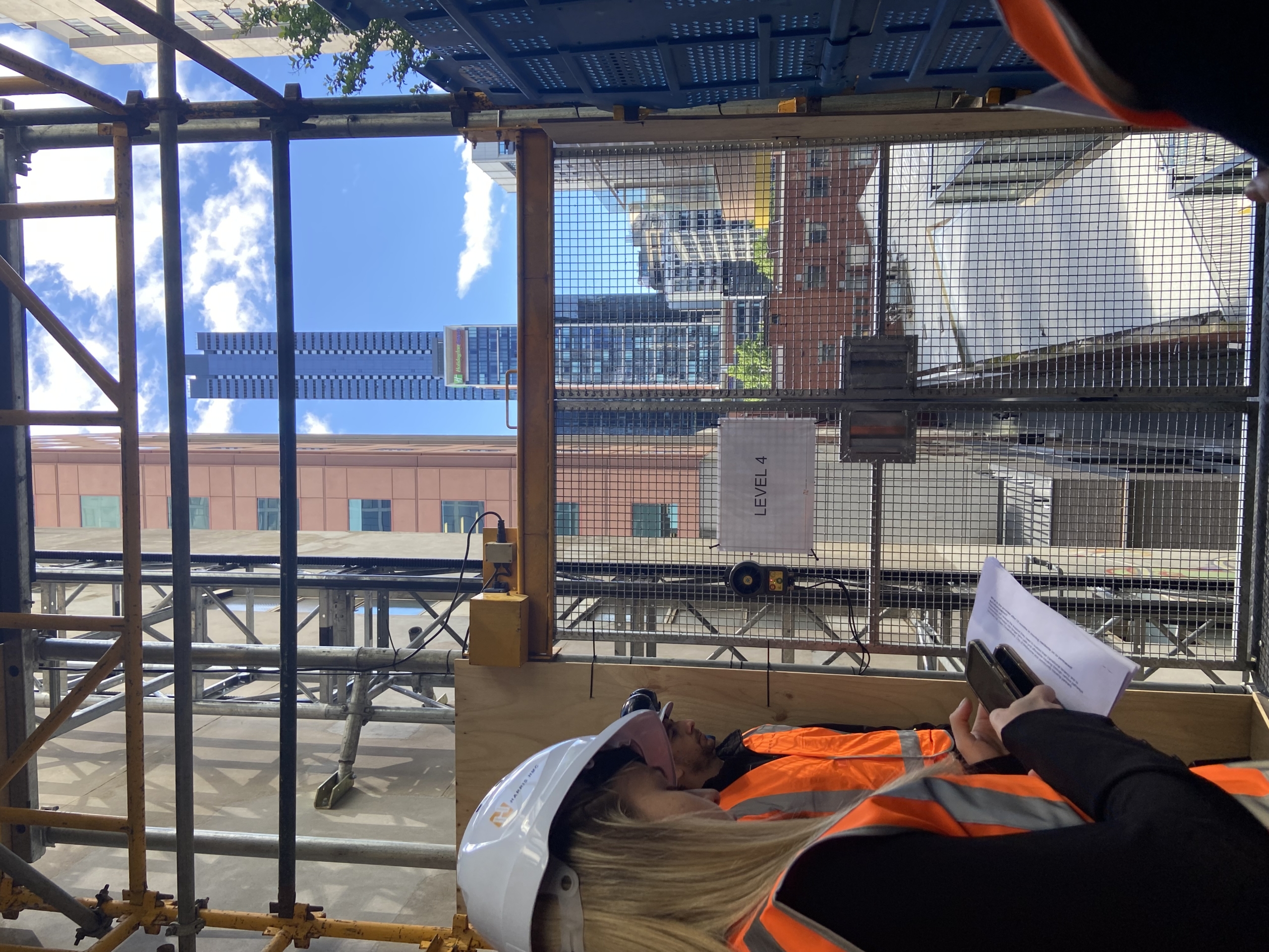 People in safety vests and hard hats stand on a construction site at Level 4, with city skyscrapers and a blue sky visible through the scaffolding in the background. One person holds papers and a tablet.