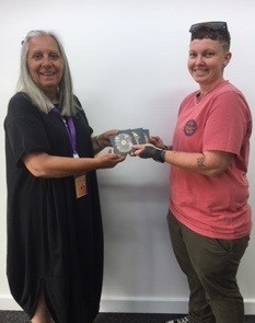 Two people stand facing each other and smiling. One, dressed in a black dress, hands over a small round object in a case to the other, who is wearing a pink shirt and khaki pants. They are in front of a plain white wall.