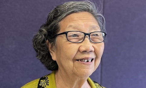 An elderly woman with short gray hair and glasses smiles, wearing a yellow sweater with a black embroidered collar and a sparkly yellow-striped shirt, standing in front of a plain purple background.