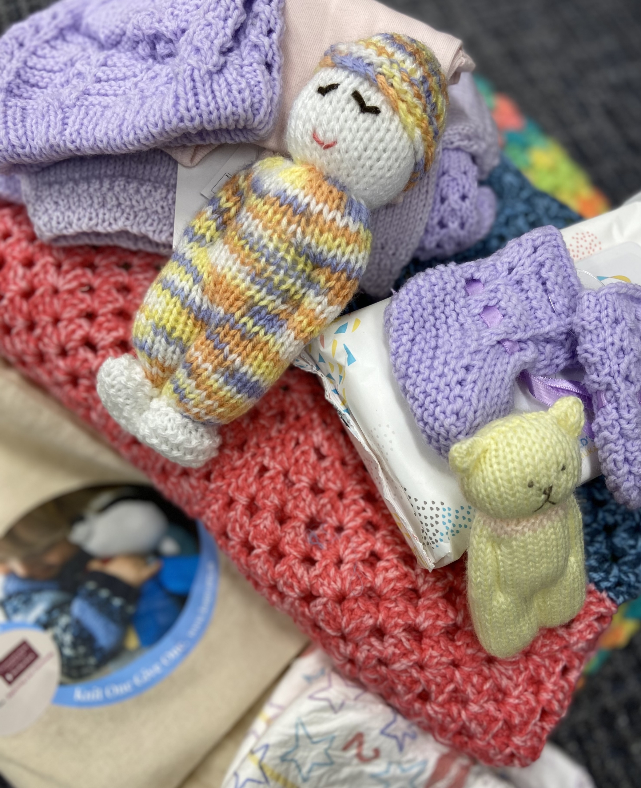 A close-up of a pile of handmade baby items including a multicolored crocheted doll, a small yellow knitted bear, purple and pink knit clothing, and a red crocheted blanket on a patterned surface.