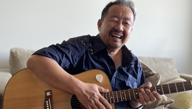 A man with short dark hair and a goatee smiles while playing an acoustic guitar, sitting on a light-colored couch in a bright room.