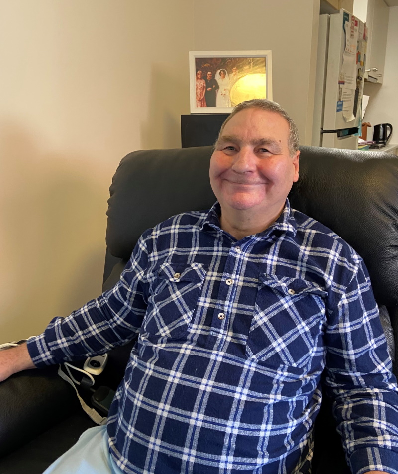 A smiling older man wearing a blue and white plaid shirt sits in a black recliner. Behind him is a framed family photo, some appliances, and a fridge in a cozy indoor setting.