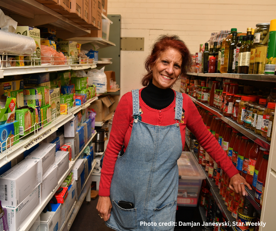 A woman with red hair, wearing a red long-sleeve shirt and denim overalls, smiles while standing in a pantry filled with food items such as pasta, sauces, and boxed goods. Photo credit: Damjan Janesvski, Star Weekly.
