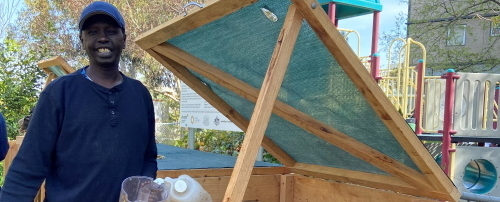 A person stands next to an open wooden compost bin outdoors, holding a plastic jug. Playground equipment and trees are visible in the background.