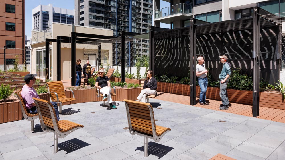 People sit and stand on a modern rooftop patio with wooden benches, planters, and pergolas, surrounded by tall buildings under a sunny sky. Some are chatting while others relax and enjoy the space.