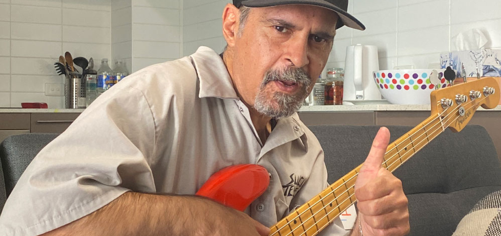 A man with a goatee and cap sits indoors on a couch, playing a red electric bass guitar and giving a thumbs up. The background shows a modern kitchen with utensils and colorful bowls.