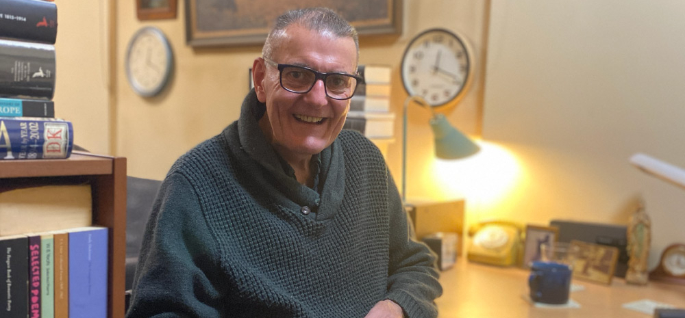 A smiling older man wearing glasses and a dark sweater sits at a desk with books, a lamp, and clocks in a warmly lit office.
