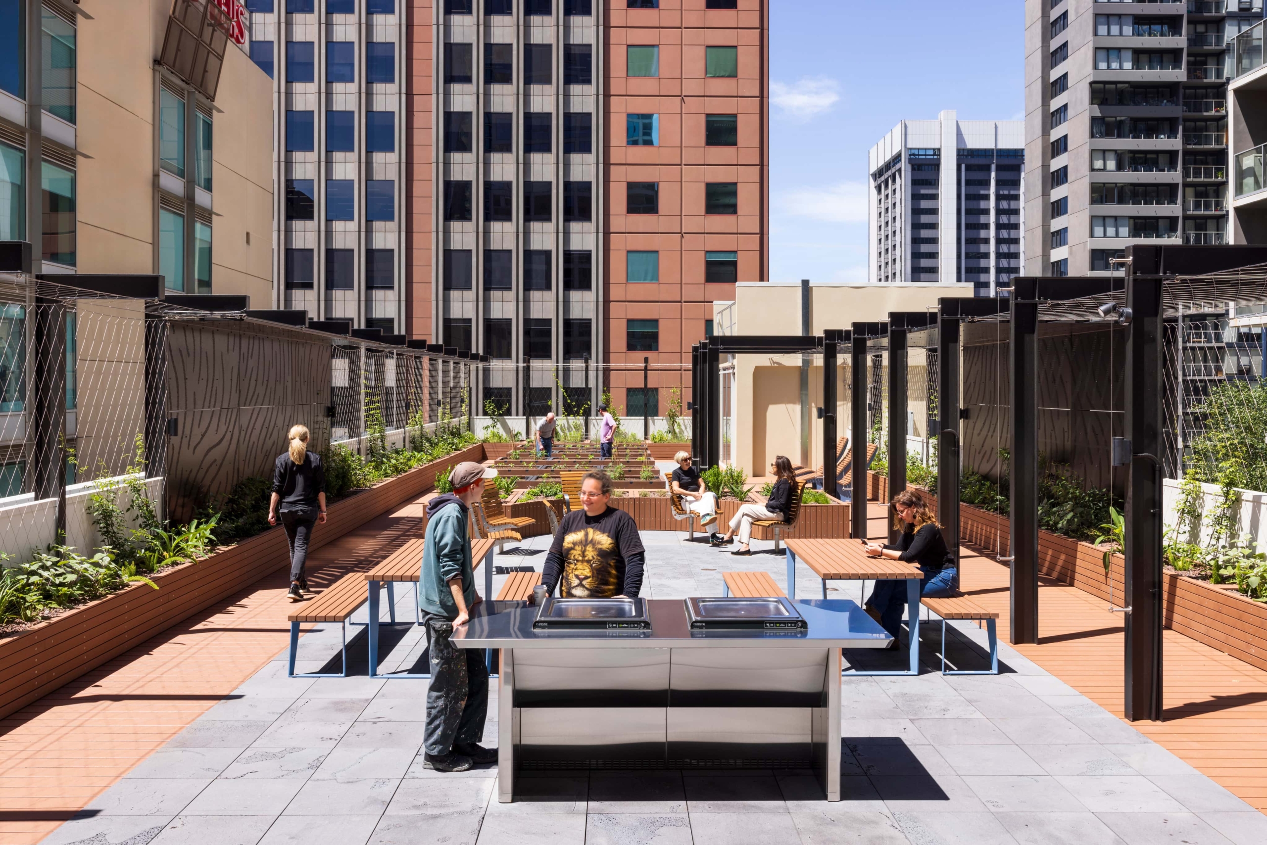 People socialize and relax on a modern rooftop terrace with benches, greenery, and pergolas, surrounded by tall office buildings under a sunny blue sky. Two men stand near a stainless steel BBQ grill in the foreground.