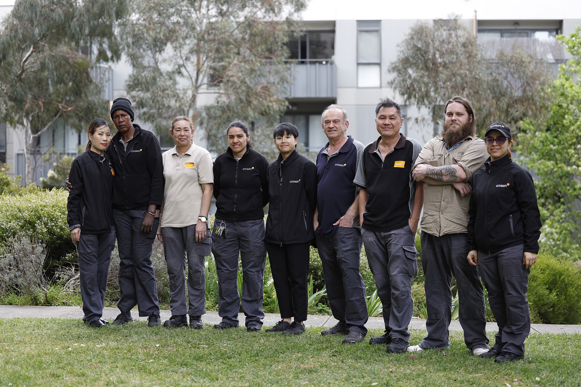 A group of nine people wearing work uniforms standing together outside on grass, with greenery and modern apartment buildings in the background. They are posing and smiling for the photo.