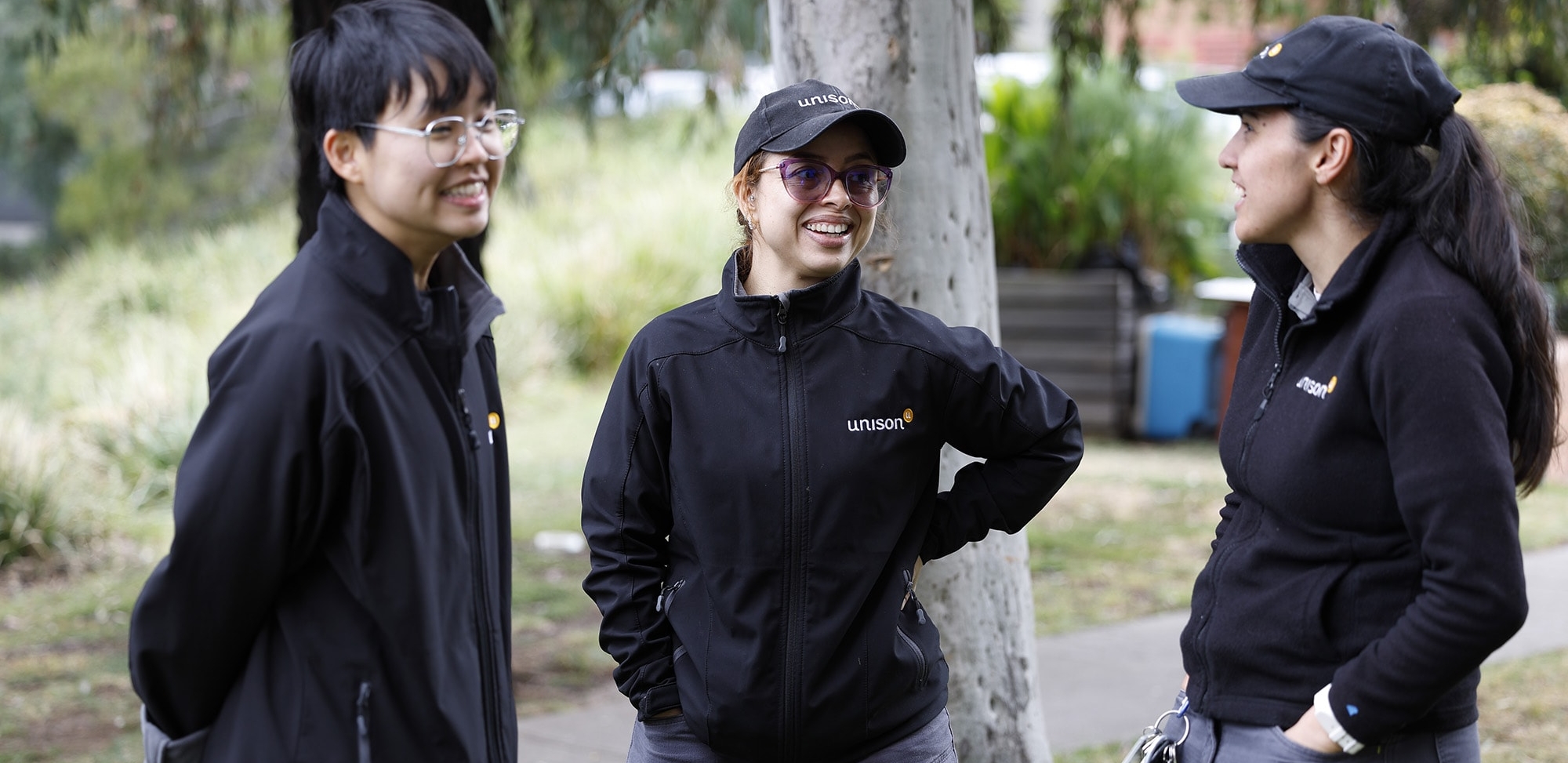 Three people wearing black Unison jackets and hats are standing outdoors, smiling and talking. They appear to be engaged in a friendly conversation, with trees and greenery in the background.