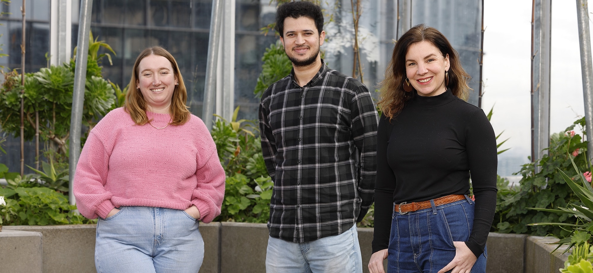 Three people standing together outdoors, smiling at the camera. There is greenery and glass structures behind them. The person on the left wears a pink sweater, the middle person wears a black plaid shirt, and the right person wears a black top.