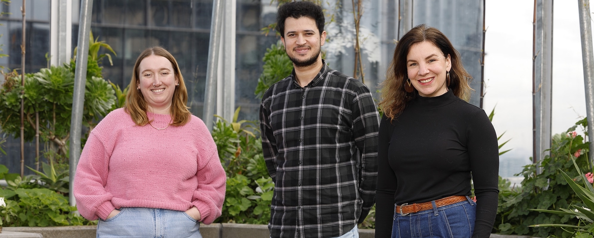 Three people standing together outdoors, smiling at the camera. There is greenery and glass structures behind them. The person on the left wears a pink sweater, the middle person wears a black plaid shirt, and the right person wears a black top.