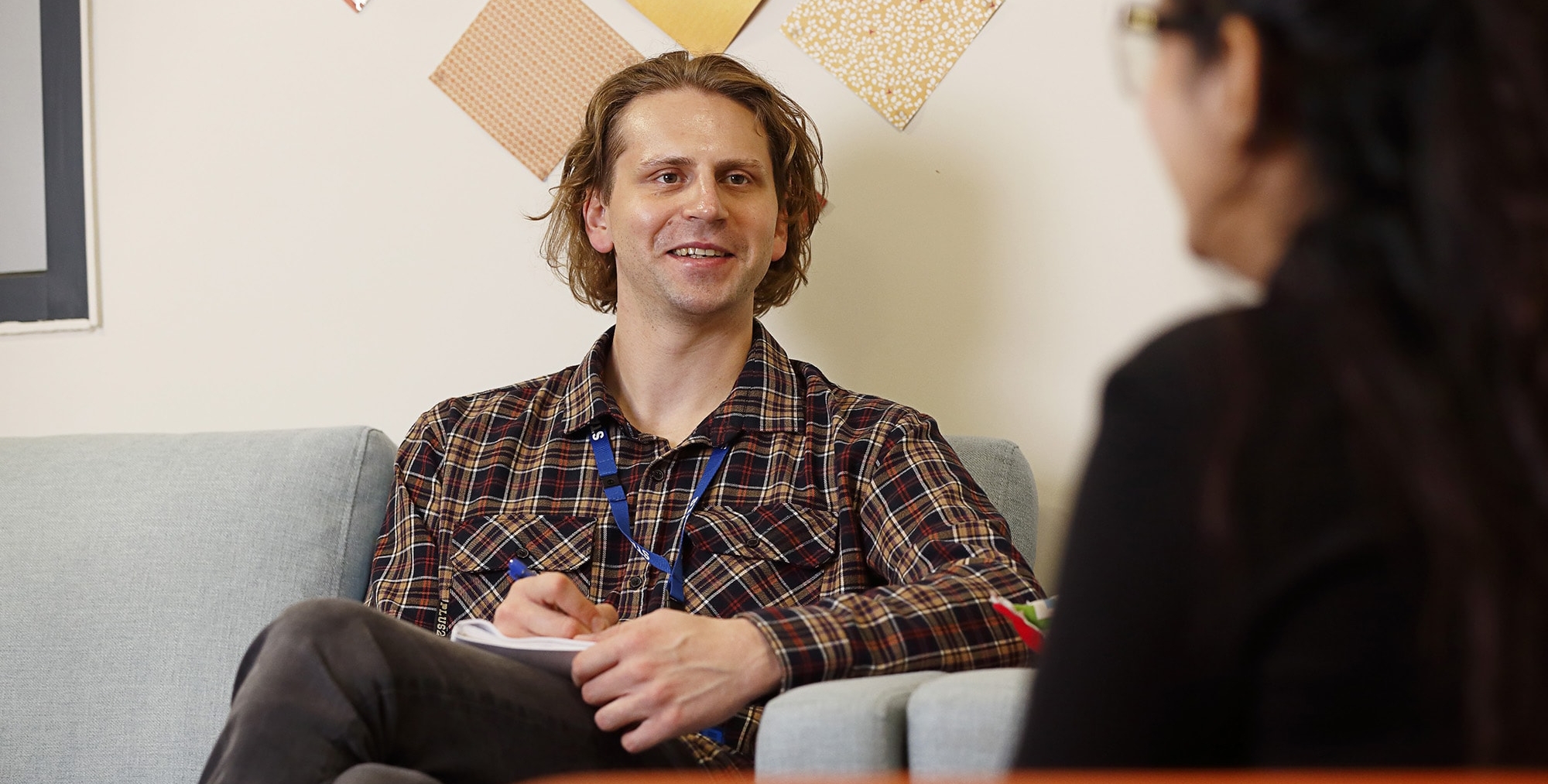 A man with shoulder-length hair wearing a plaid shirt sits on a couch, smiling and holding a notepad while talking to a woman with dark hair who is seated across from him.