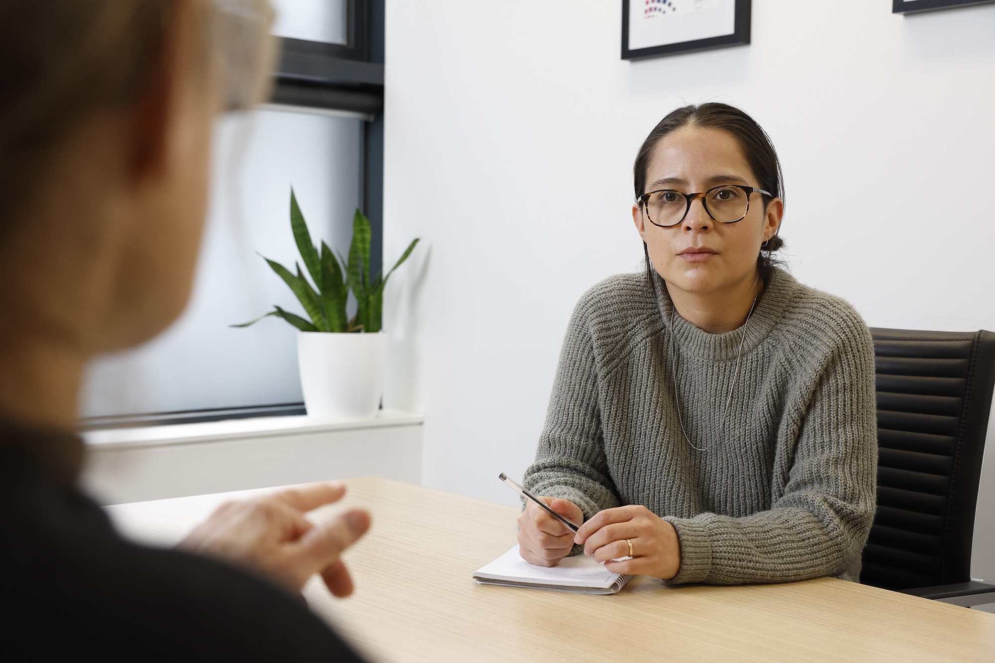 A person wearing glasses and a gray sweater sits at a desk, holding a pencil and notepad, listening attentively to another person whose back is turned. A potted plant sits on the windowsill in the background.