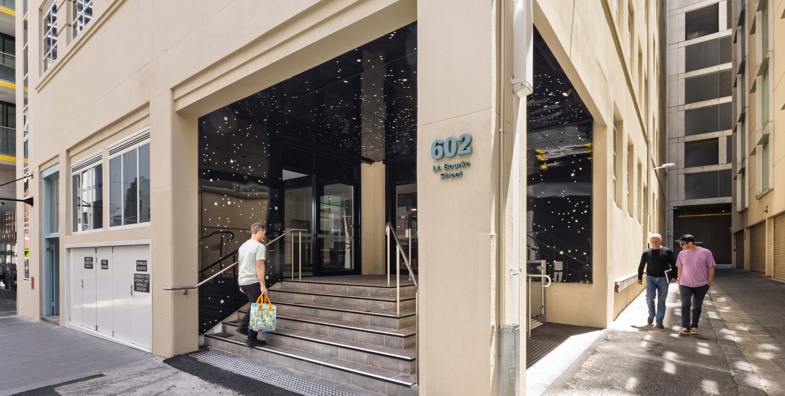 Modern building entrance at 602 L.A. Reynolds Square with stairs, glass doors, and a man carrying a colorful bag ascending. Two people walk past on the adjacent sidewalk in an urban setting.