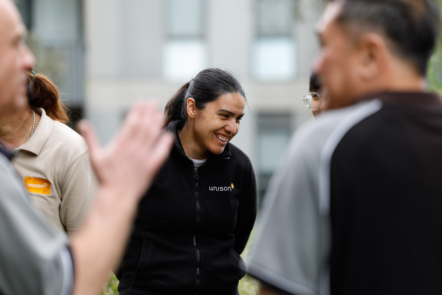 A woman wearing a black Unison jacket smiles while standing outdoors with a group of people, some of whom wear grey or beige Unison shirts. The background is blurred.