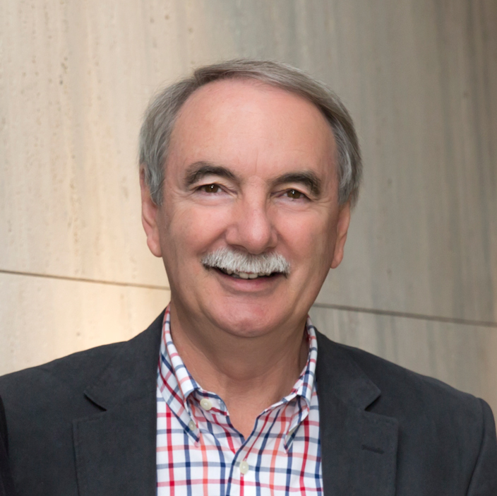 A smiling older man with gray hair and a mustache, wearing a checked shirt and dark blazer, stands in front of a light-colored, textured wall.