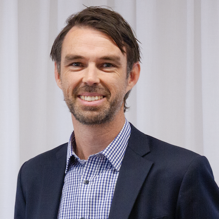 A man with short brown hair, a beard, and a friendly smile is wearing a navy blazer over a checkered shirt, standing in front of white curtains.
