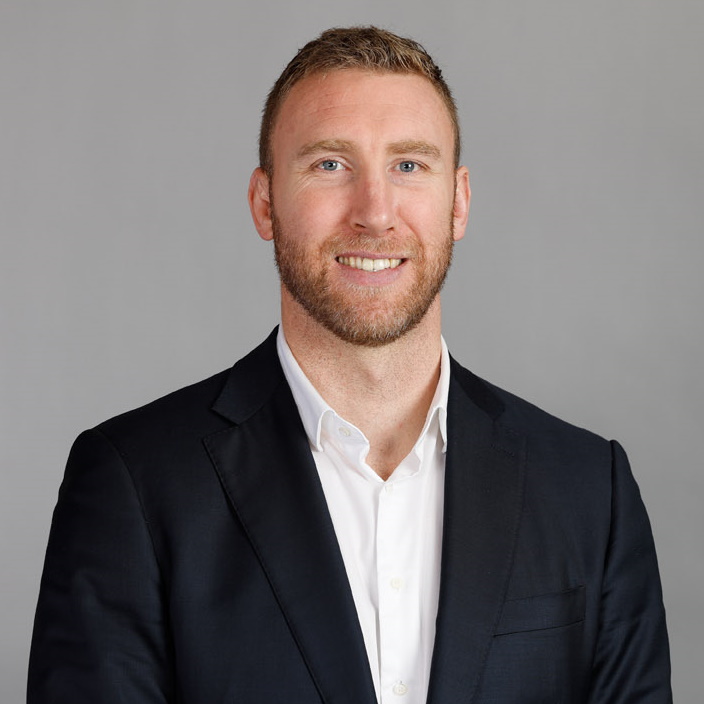 A man with short light brown hair and a beard, wearing a white dress shirt and a dark blazer, smiles at the camera against a plain gray background.