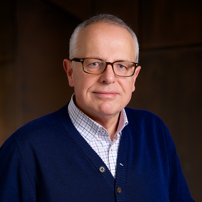 A middle-aged man with short gray hair and glasses, wearing a blue sweater over a white checkered shirt, smiles gently in front of a dark, blurred background.