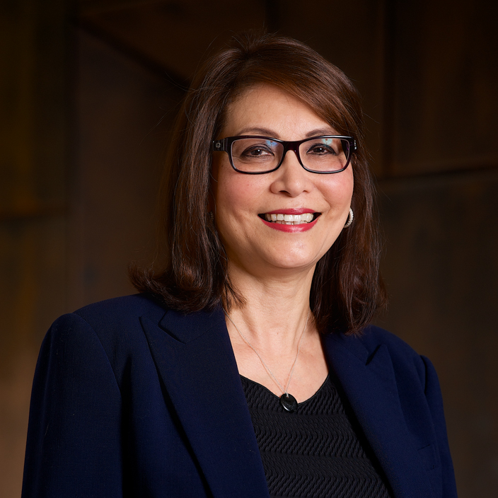 A woman with shoulder-length brown hair and glasses, wearing a navy blazer over a black top, smiles confidently at the camera against a dark, blurred background.