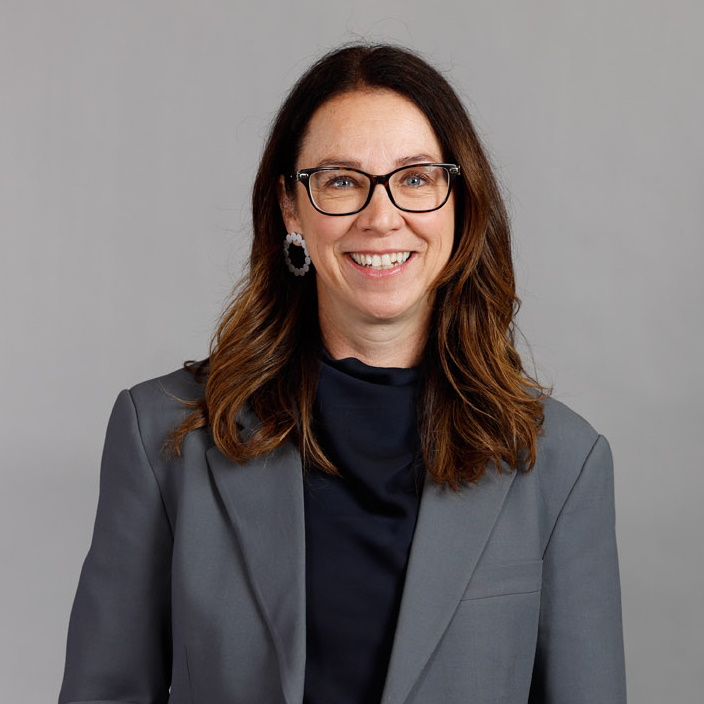 A woman with long brown hair, wearing glasses, a gray blazer, and a dark top, smiles in front of a plain gray background.