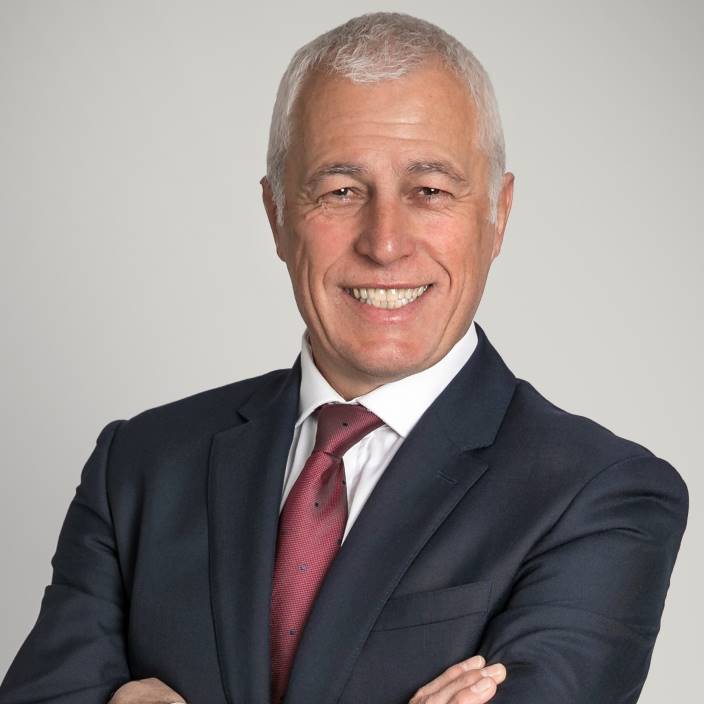 Smiling older man with short gray hair in a dark suit, white shirt, and red tie, standing with arms crossed against a plain light background.