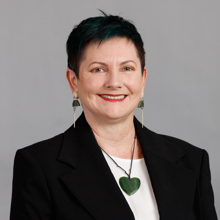 A woman with short dark hair and green highlights, wearing a black blazer, white top, green heart necklace, and green earrings, smiles in front of a plain gray background.