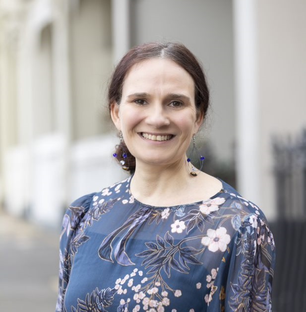 A woman with dark hair tied back, wearing a blue floral blouse and drop earrings, smiles while standing outdoors on a city street with blurred buildings in the background.
