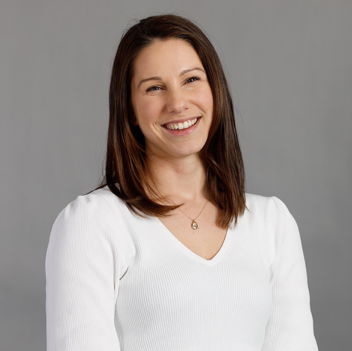 A woman with straight brown hair, wearing a white long-sleeve top and a necklace, smiles while sitting against a plain gray background.