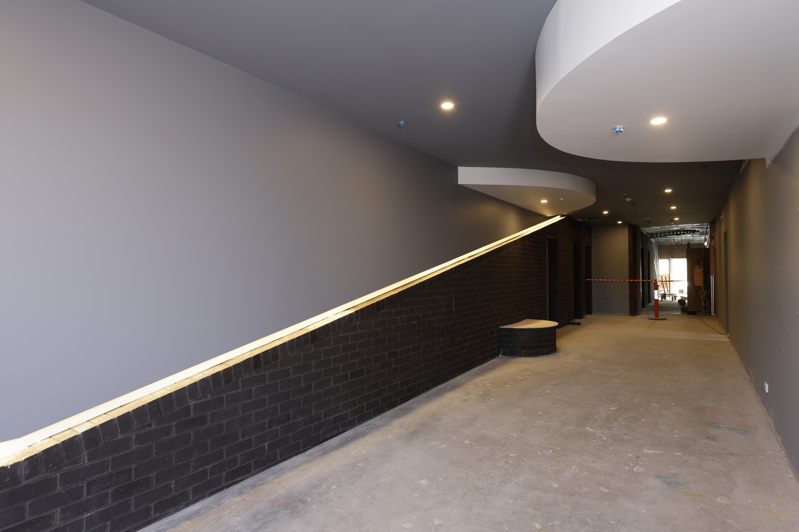 A modern hallway under construction with gray walls, ceiling lights, black brick lower walls, and a curved white ceiling feature. The floor is unfinished, and the far end is blocked with caution tape and orange cones.