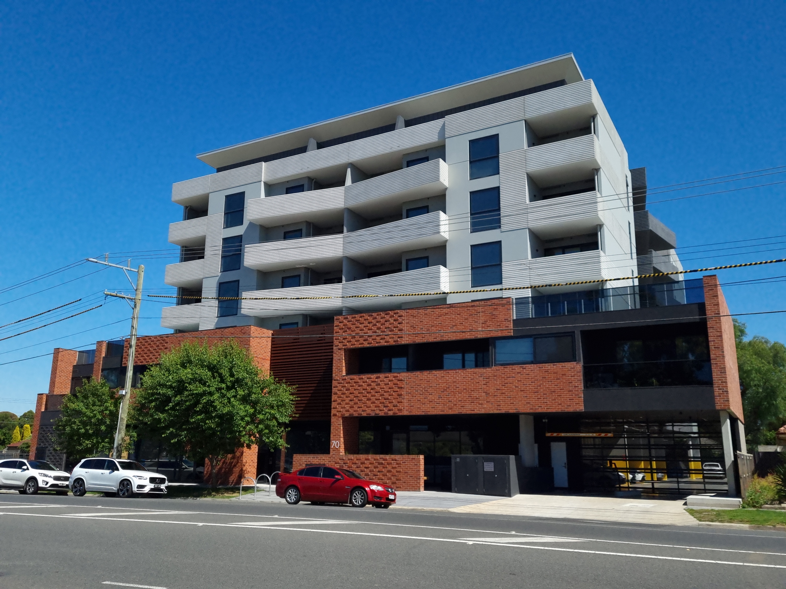 A modern six-story apartment building with red brick and gray panels, featuring multiple balconies. Several cars are parked along the street in front under a clear blue sky.