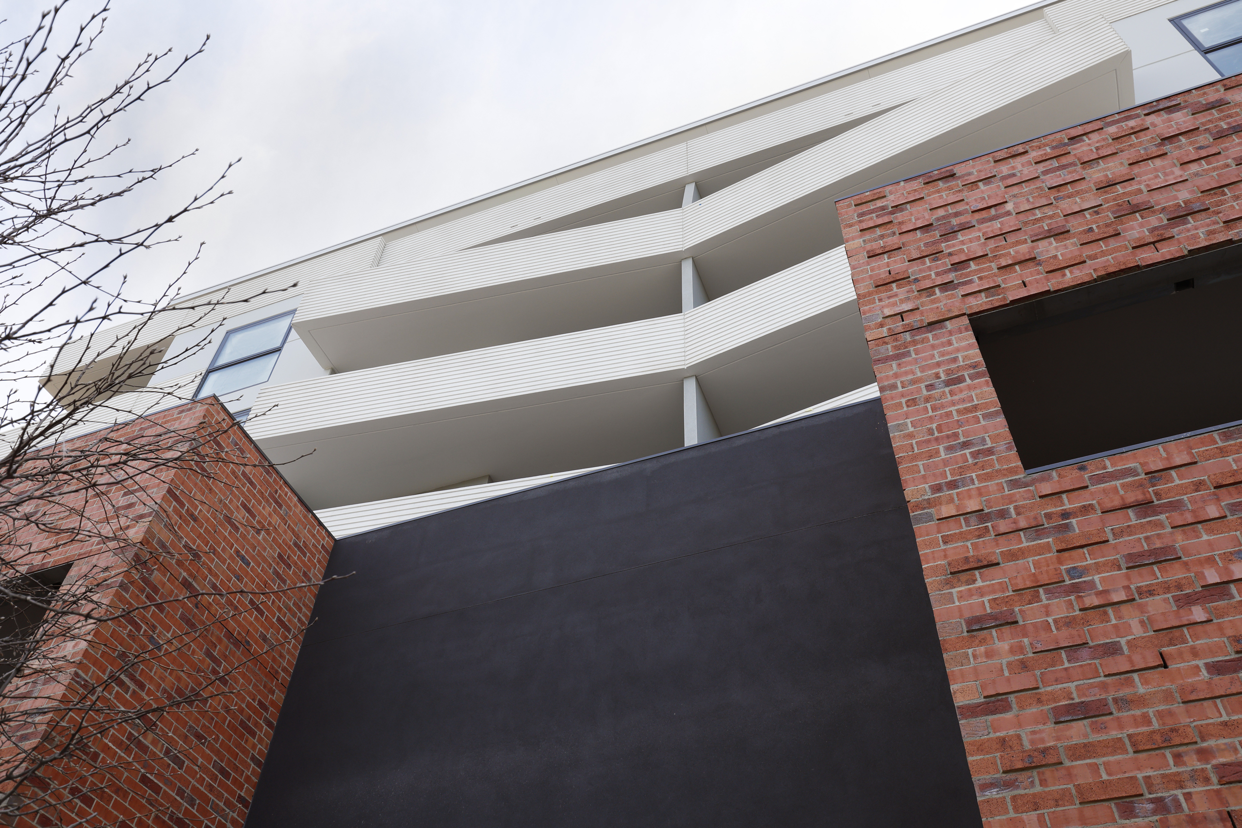 Low-angle view of a modern apartment building with white balconies, reddish-brown brick walls, a black central section, and a leafless tree on the left against a cloudy sky.