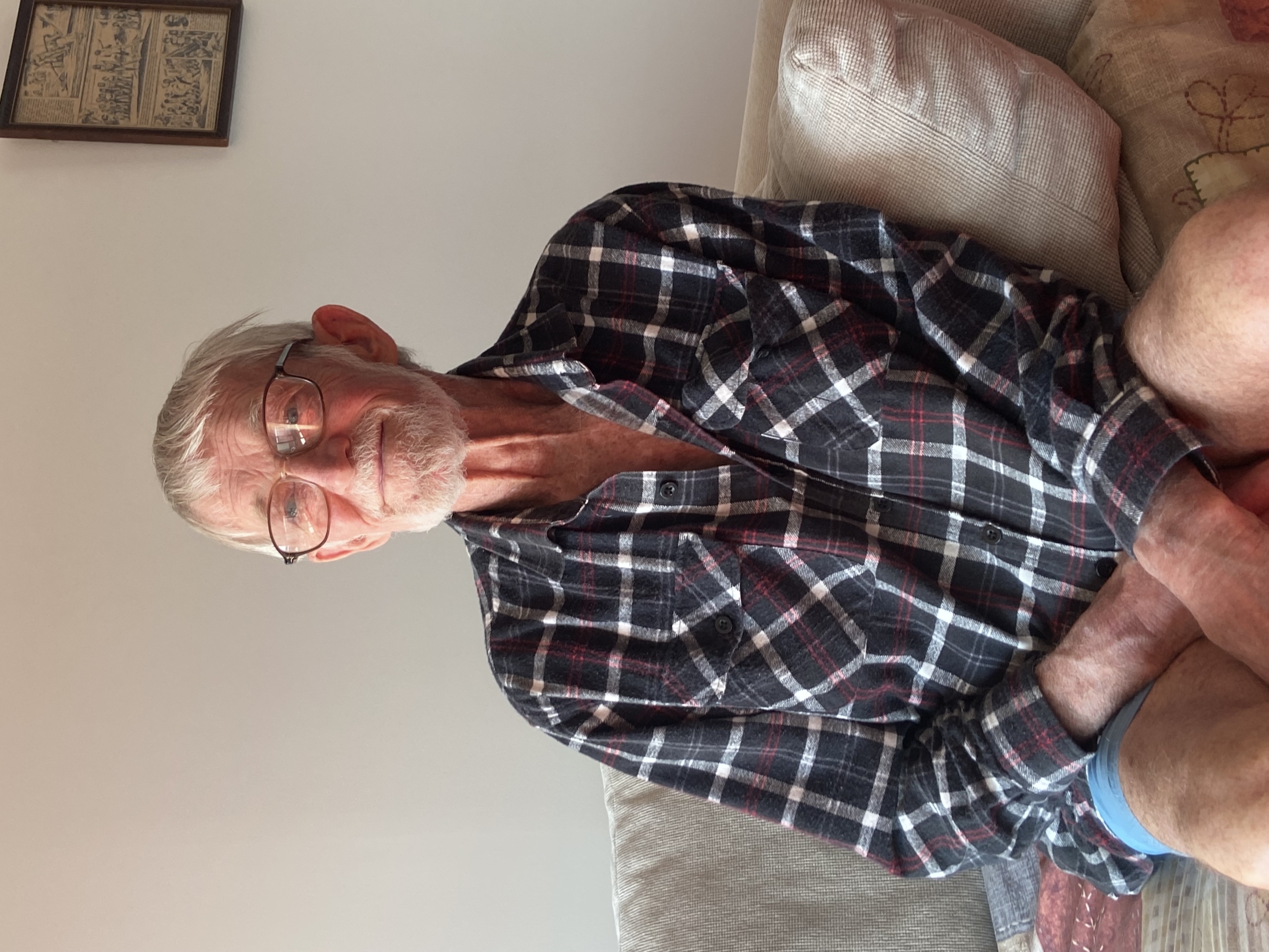 An elderly man with white hair, glasses, and a beard sits on a beige couch, wearing a black and red plaid shirt and shorts, smiling slightly. A framed picture hangs on the wall behind him.