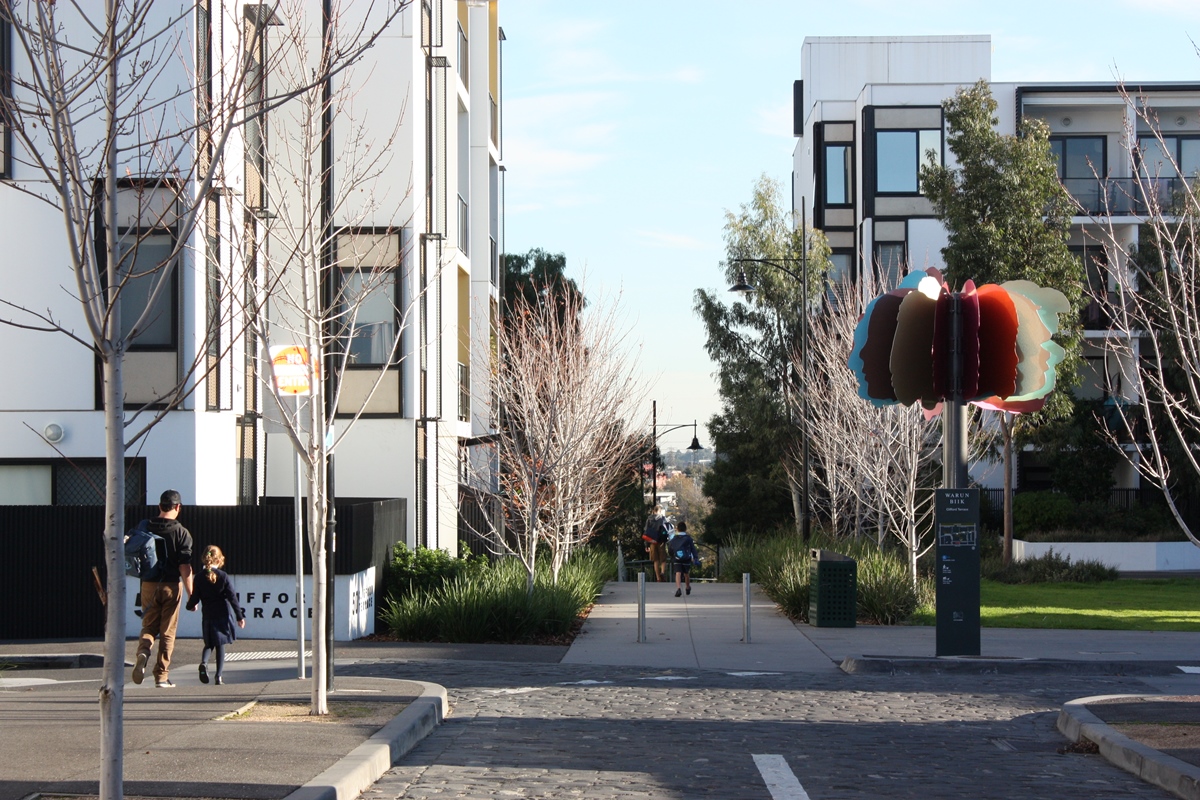 A man and child walk on a footpath between modern apartment buildings, with bare trees and a colorful abstract sculpture on a pole nearby. It is a sunny day.