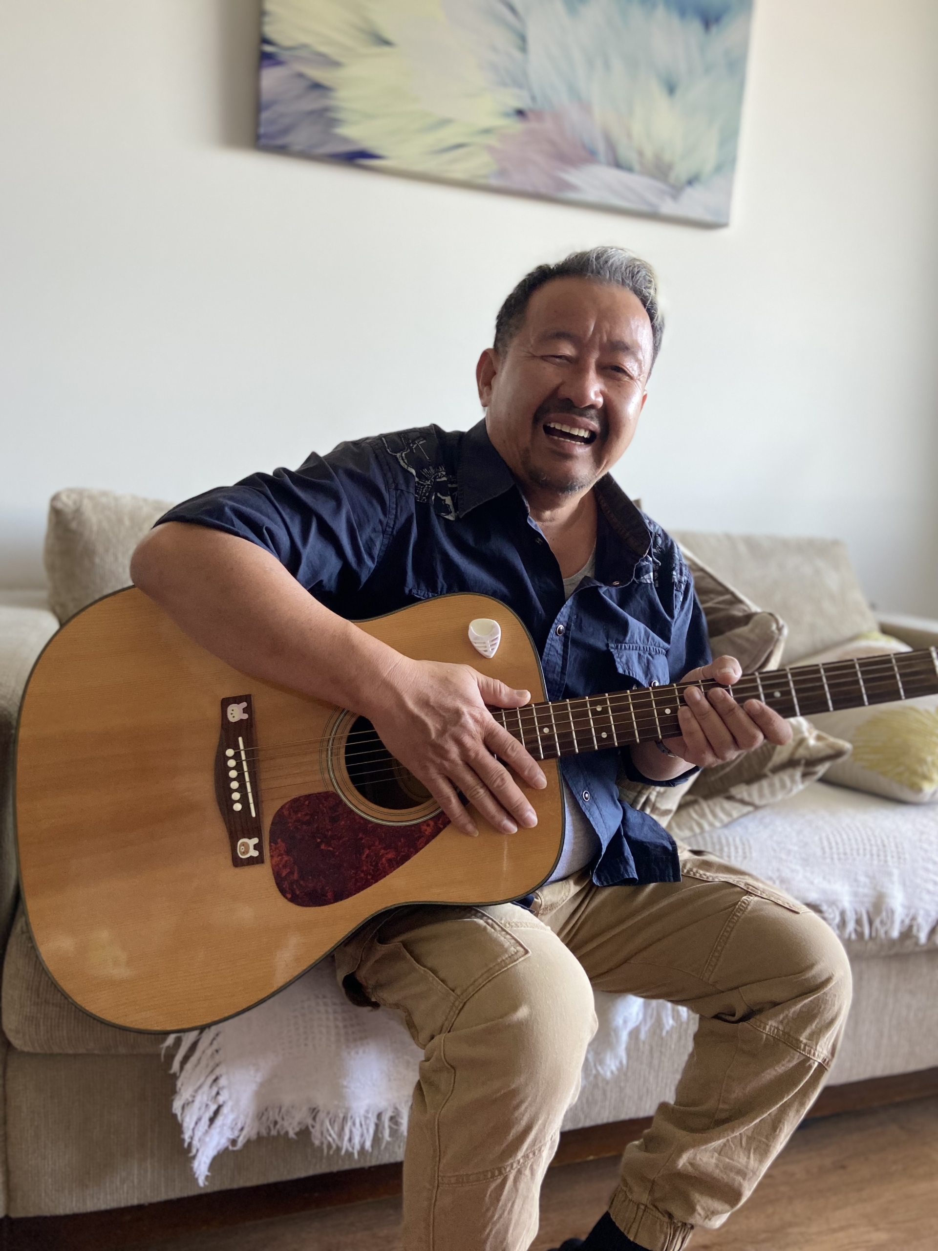 A smiling man sits on a couch playing an acoustic guitar in a cozy living room, with a colorful abstract painting hanging on the wall behind him.