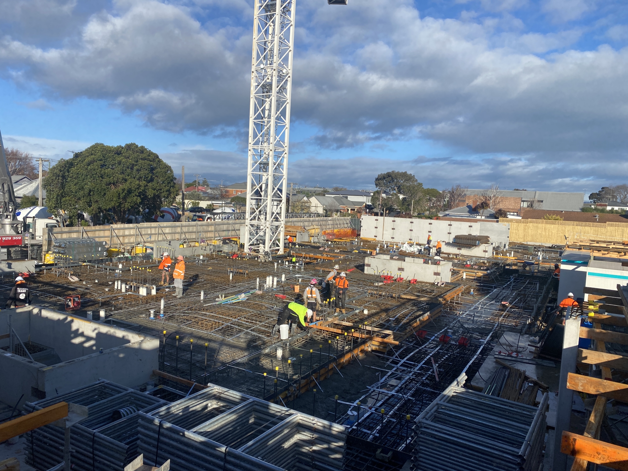Construction site with workers in safety vests and helmets building a large structure. Steel rebar, pipes, and a tall crane are visible under a partly cloudy sky, with trees and buildings in the background.